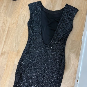 Svart glittrig paljettklänning - Säljer en svart kort klänning med glittriga paljetter över hela ytan. Klänningen har djup v-ringning och är ärmlös, vilket ger en snygg och festlig look. Materialet är täckt av paljetter som reflekterar ljuset och gör dig till kvällens stjärna.
