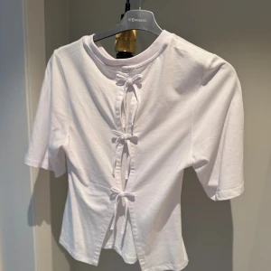 Vit topp med snörning i ryggen - Vit kortärmad topp i bomull med rund hals och snygg snörningsdetalj i ryggen. Toppens passform är normal och den har en stilren look framtill medan ryggen sticker ut med tre knytband som ger en cool twist.