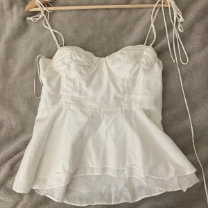Vit linnetopp från Zara med snörning - Supersöt vit linnetopp från Zara med tunna axelband som knyts och markerade kupor. Toppen har en lätt utsvängd nederdel och smock i ryggen för extra komfort. Perfekt för varma dagar och enkel att styla med jeans eller kjol.