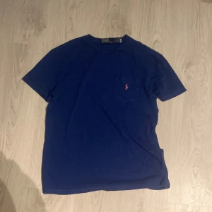 Blå t-shirt från Polo Ralph Lauren - Snygg marinblå t-shirt från Polo Ralph Lauren med klassisk passform. T-shirten har en liten ficka på bröstet med rosa broderad logga. Perfekt för dig som gillar stilrena och enkla plagg med en touch av ikonisk design.