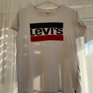 Vit t-shirt med Levis-logga - Vit t-shirt från Levi's med klassisk stor logga i svart och rött framtill. T-shirten har rund halsringning och korta ärmar. Tillverkad i mjuk bomull, perfekt för en avslappnad och stilren look.