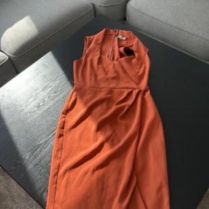 Rostfärgad omlottklänning från ASOS - Säljer en snygg rostfärgad omlottklänning från ASOS med djup v-ringning och ärmlös design. Klänningen har en draperad front och dold dragkedja i ryggen. Perfekt för dig som gillar stilrena och moderna plagg med lite edge.