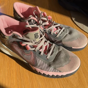 Nike Kyrie basketskor i grått och rosa - Snygga Nike Kyrie basketskor med grå mesh, rosa detaljer och camo-mönstrade partier på hälen. Skorna har snörning, rund tå och svart sula med rosa logga undertill. Kyrie Irving känd för att göra snabba, rappa skor med bra grepp och stabil mobilitet
