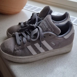 Grå adidas Campus sneakers i mocka - Snygga grå adidas Campus sneakers med klassiska vita ränder på sidorna. Perfekta för dig som gillar en clean och tidlös streetstil. Knappt använda då de är för små för mig:(
