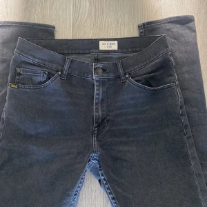 Mörkgrå jeans från Tiger of Sweden - Snygga mörkgrå jeans från Tiger of Sweden med klassisk femficksdesign och diskret logga på bakfickan. Jeansen har raka ben och normal passform, perfekta till en avslappnad stil. Tillverkade i mjukt denimtyg för skön känsla hela dagen.