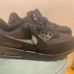 Nike Air Max 90 Triple Black - Nike Air Max 90 Triple Black med ikonisk synlig Air-enhet i sulan. Hela skon är svart med en mix av skinn och syntetmaterial, klassisk swoosh på sidan och snörning. Perfekt för dig som gillar stilrena och sportiga sneakers.