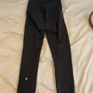 Svarta leggings från Lululemon - Svarta, tighta leggings från Lululemon med hög midja och klassisk logga vid benet. Perfekta för träning eller chill, tillverkade i ett mjukt och stretchigt material som sitter skönt hela dagen.