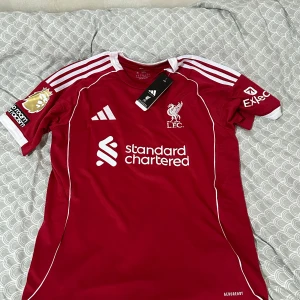 Liverpool FC röd matchtröja Adidas - Liverpool FCs officiella matchtröja i rött med vita detaljer, tillverkad i lätt och ventilerande polyester. Adidas-logga på bröstet, klubbmärke broderat och sponsortryck framtill. Nummer 7 och namnet Wirtz på ryggen. Klassiska vita ränder på axlarna. Är is storlek M men passar S också Pris kan diskuteras!