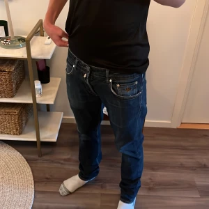 Mörkblå Morrisjeans med broderad ficka - KÖPTA FÖR 1599 SÄLJS FÖR 750! Säljer ett par mörkblå jeansbyxor med snygga slitningar och en broderad detalj på myntfickan. Klassisk femficksmodell med raka ben och normal passform. Perfekta för dig som gillar en avslappnad stil med lite extra detaljer.