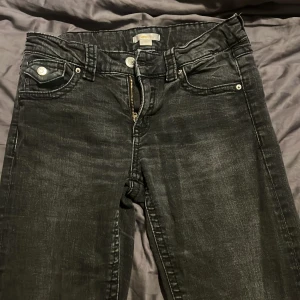 Svarta bootcut jeans med fickdetaljer - Snygga svarta jeans med lätt tvättad look och bootcut passform. Byxorna har fram- och bakfickor med lock och silverfärgade knappar. Klassisk dragkedja och bälteshällor. Perfekta för dig som gillar en stilren men edgy vibe.