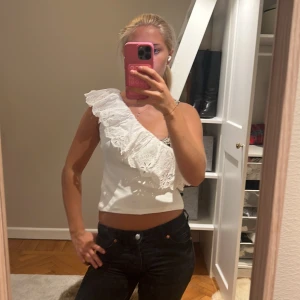 Vit oneshoulder topp med volang från zara - Säljer en vit croppad topp med oneshoulder-design och stor volang med broderade blommor. Toppen är ärmlös på ena sidan och har en snygg asymmetrisk look som sticker ut. Perfekt för dig som gillar unika detaljer och somrig stil. Den är som storlek xs