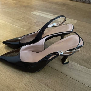 Bruna lackpumps med spetsig tå - Snygga bruna pumps i lack med spetsig tå och öppen häl. Skorna har en elegant låg klack och justerbart spänne i guld. Perfekta för dig som vill ha en stilren och trendig look.