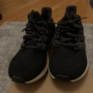 Svarta Adidas Ultraboost sneakers - Svarta Adidas Ultraboost sneakers med stickad ovandel och klassiska tre ränder i plast på sidorna. Skorna har snörning och en vit, responsiv Boost-sula för extra komfort. Perfekta för dig som gillar stilrena och sportiga skor med modern känsla. Storlek 40 men passar mer 41