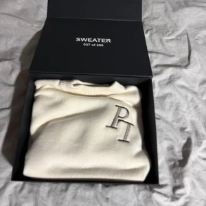 Vit stickad tröja från Penelope - Exklusiv (037 av 200) vit stickad tröja från Penelope med broderad PL-logga på bröstet. Tröjan har ribbade muddar och nederkant, rund halsringning och en clean, minimalistisk look. Perfekt för dig som gillar stilrena vibes och limited edition-plagg.