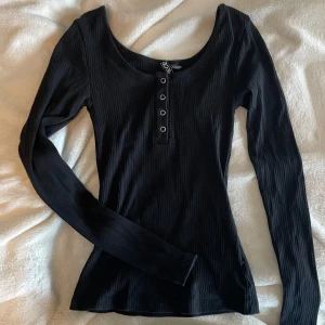 Svart långärmad topp - Svart ribbad långärmad topp från HM med rundad halsringning och fyra dekorativa knappar framtill. Snygg slim passform och stretchigt material som sitter skönt på kroppen. Perfekt att styla med jeans eller kjol för en clean look.