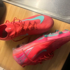 Nike Mercurial rosa fotbollsskor - (Nypris CA 3000)(Snygga Nike Mercurial fotbollsskor i en stark rosa färg med ljusblå swoosh och detaljer. Skorna har snörning, platt sula med dobbar och är tillverkade i ett lätt syntetmaterial som andas. Perfekta för dig som vill sticka ut på planen.