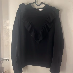 Svart blus  - Svart blus från H&M i storlek 42 med lång ärm och snygg volangdetalj framtill som ger en cool och trendig look. Blusen har rund hals och är gjord i ett mjukt, lätt material som känns skönt mot huden. Perfekt för dig som gillar stilrena plagg med en twist.