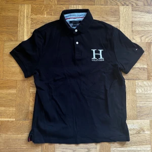 Svart Tommy Hilfiger polo t-shirt - Svart polo t-shirt från Tommy Hilfiger med stor H-brodyr och logga på bröstet. Klassisk krage med två knappar och custom fit-passform. Perfekt för en clean och stilren look.