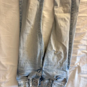 Bootcut ljusblå jeans från H&M - Säljer ett par ljusblå bootcut jeans från H&M med låg midja och justerbar midja. Jeansen har klassiska bakfickor med lock och knappar, samt snygg tvättad denimlook. Perfekta för dig som gillar en avslappnad och trendig stil.