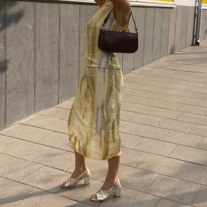 Gul melerad långklänning i satin  - Säljer en flowy långklänning i satin med ett abstrakt mönster i gula och beige toner. Klänningen är ärmlös och har en rund halsringning, perfekt för varma dagar. Materialet har en lätt glans och ger en lyxig känsla. Passformen är normal och klänningen faller snyggt längs kroppen.