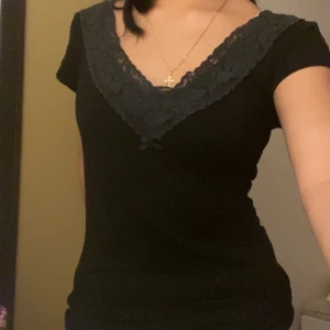 Svart blus med spets - Svart blus från Lindex med v-ringning och bred spetskant framtill. Toppen har korta ärmar och en liten rosett vid urringningen. Snygg och enkel modell som passar till många olika looks.