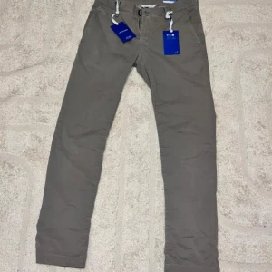 Grå chinos från Jacob Cohën, stl 32 - Snygga grå chinos från Jacob Cohën med raka ben och klassisk passform. Byxorna har diskret logopatch bak, dragkedja och knäppning fram samt fickor både fram och bak. Tillverkade i mjuk bomull för en clean och stilren look.