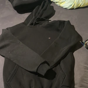 Svart hoodie från Tommy Hilfiger - Säljer en svart hoodie från Tommy Hilfiger med liten broderad logga på bröstet. Tröjan har huva med snörning och en stor magficka. Perfekt för dig som gillar en enkel och stilren look.