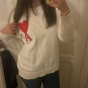 Vit stickad tröja med rött hjärta - Säljer en vit stickad tröja från AMI Paris med ett stort rött hjärta och bokstaven A broderat på bröstet. Tröjan har lång ärm, ribbade muddar och en avslappnad passform. 
