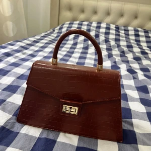 Brun handväska i croco-mönster - Säljer en snygg brun handväska med croco-mönster och guldfärgade detaljer. Väskan har ett stilrent handtag, vridlås framtill och dragkedja på baksidan. Perfekt accessoar för att lyfta din outfit.