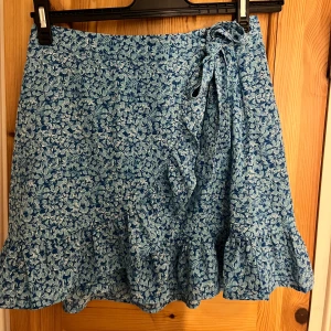 Blommig blå omlottkjol med volang - Supersöt kort omlottkjol i blått med blommigt mönster och volangkant. Kjolen har knytband i midjan.