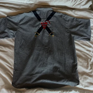 Rutig skjorta med broderade hundar Brioche - Unik Disney inspirerad kortärmad skjorta från Brioche i svartvitt rutigt mönster. På framsidan finns broderade dalmatiner och detaljer som liknar hängslen, med texten 'Happy Top' på ryggen. 