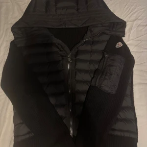 Moncler cardigan  - Svart pufferjacka från Moncler med stickade ärmar och huva. Jackan har dragkedja framtill, en ficka med dragkedja på ärmen och ikonisk Moncler-logga på axeln. Kombinerar quiltad pufferstil med stickade detaljer för en modern look de är bara att höra av sig för fler frågor och bilder🙌