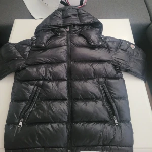 Moncler maya jacka - Svart pufferjacka från Moncler med glansig finish, huva och två dragkedjeförsedda fickor. Jackan har klassisk Moncler-logga på ärmen och är fylld med certifierat dun. Perfekt för kalla dagar. Storlek 3 som motsvarar L
