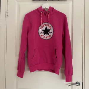 Rosa hoodie från Converse - Säljer en snygg rosa hoodie från Converse med stort broderat All Star-märke på bröstet. Tröjan har huva med snörning, magficka och långa ärmar. Perfekt för dig som gillar streetstyle och vill ha något bekvämt och färgglatt.