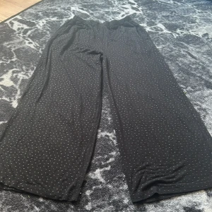 Svarta prickiga wide byxor - Säljer ett par svarta byxor med små prickar och breda ben. Byxorna har elastisk midja med justerbar knappresår och en bakficka med en liten gulddetalj. Perfekta för en chill och avslappnad stil.