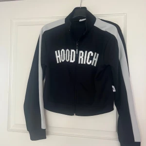 Svart ziptröja från Hoodrich - Svart ziptröja från Hoodrich med vita bokstäver på bröstet och gråa paneler längs ärmarna. Hög krage och hel dragkedja framtill. Perfekt för en sportig och streetinspirerad stil.