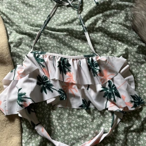 bikinitopp - Supersöt bikini med volang och palmmönster i grönt och ljusrosa på vit botten. Bikinin har knyt i nacken och volanger framtill som ger en trendig och somrig look. Perfekt för strandhäng och poolparty!