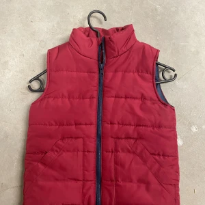 Röd quiltad dunväst från H&M - Snygg röd dunväst från H&M med quiltad design och hög krage. Västen har två snedställda fickor framtill och stängs med en mörk dragkedja. Perfekt för lager-på-lager under kyliga dagar.