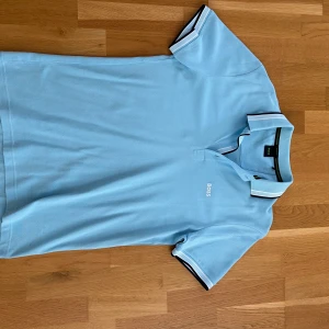 Ljusblå BOSS pikétröja Regular Fit - Snygg ljusblå pikétröja från BOSS med klassisk krage och broderad logga på bröstet. Tröjan har korta ärmar och vita detaljer på krage och ärmslut. Tillverkad i bomull för en skön och luftig känsla. Perfekt för en clean och stilren look. Storlek M 