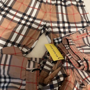 Rutig Burberry shorts med snörning - Snygga shorts från Burberry i klassiskt beige, svart, vit och röd rutig design. De har elastisk midja med svart snörning och är tillverkade i ett lätt syntetmaterial. Perfekta för sommaren och ger en trendig vibe till din outfit. Som är i storlek S/M pris 450 kr 