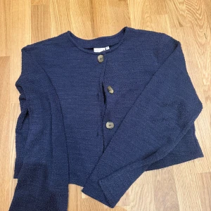 Mörkblå kofta från Nelly med knappar - Mysig mörkblå kofta från Nelly i storlek XS. Koftan har en rak passform, rund halsringning och stora knappar framtill. Materialet är mjukt och lite strukturerat, perfekt för lager-på-lager under kyliga dagar.