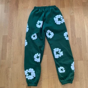 Gröna mjukisbyxor Denim Tears L - Gröna mjukisbyxor från Denim Tears i storlek L med vita blommiga tryck över hela byxorna. Byxorna har resår i midjan och vid bensluten för en skön och avslappnad passform. Perfekta för chill dagar och streetwear-vibe.