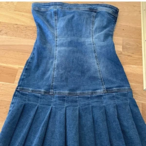 Jeansklänning från Bershka - Säljer en snygg axelbandslös jeansklänning från Bershka i storlek S. Klänningen är kort, har markerad midja och plisserad kjol. Tillverkad i klassiskt blå denim med rå kant nertill. Perfekt för dig som gillar en cool och trendig look.