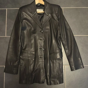 Svart skinnkavaj från Jacques - Stilren svart skinnkavaj från Jacques med klassisk krage, knäppning framtill och två stora fickor. Jackan har en rak, något längre passform och är helfodrad. Perfekt för dig som vill ha en edgy men clean look.