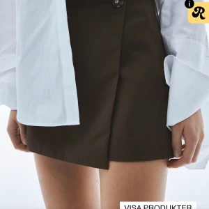 Brun omlott minikjol med knapp - Snygg brun kortkjol i omlottmodell med en stilren knappdetalj i midjan. Kjolen har en rak passform och är tillverkad i ett slätt tyg som ger en clean look. Perfekt att matcha med en oversized skjorta eller stickad tröja.