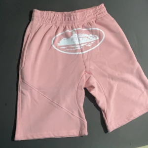 Rosa shorts från Corteiz, medium - Säljer ett par ljusrosa shorts från Corteiz. De har elastisk midja, sidofickor och en vit grafisk print framtill. Perfekta för chill dagar eller när du vill ha en avslappnad look.