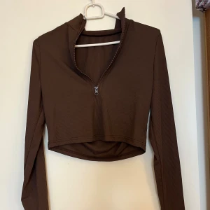 Brun croppad ribbad polotopp - Säljer en brun croppad polotopp med lång ärm och halv dragkedja framtill. Toppen är ribbad och har en tight passform som sitter snyggt. Perfekt att styla med jeans eller kjol för en trendig look.