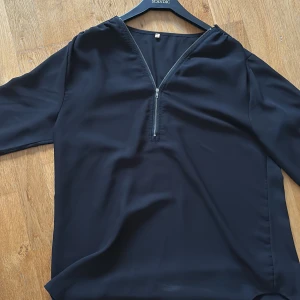 Svart blus med dragkedja - Snygg svart blus med lång ärm och v-ringning som har en silverfärgad dragkedja framtill. Blusen har en lös passform och är tillverkad i ett lätt, tunt material som ger ett stilrent intryck.