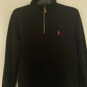 Svart stickad tröja från Ralph Lauren - Svart stickad tröja från Ralph Lauren med röd broderad logga på bröstet. Tröjan har hög krage och half zip-dragkedja framtill. Perfekt för dig som gillar klassisk och stilren design med en sportig touch.
