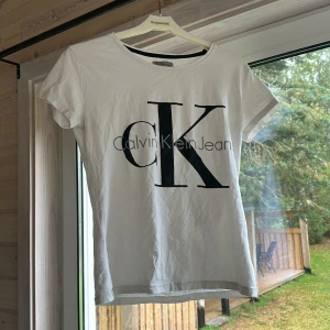 Vit t-shirt från Calvin Klein - Vit t-shirt från Calvin Klein med stor svart CK-logga och texten Calvin Klein Jeans på bröstet. Klassisk rund halsringning och korta ärmar. Tillverkad i mjuk bomull som känns skön mot huden. Perfekt för en clean och stilren look.